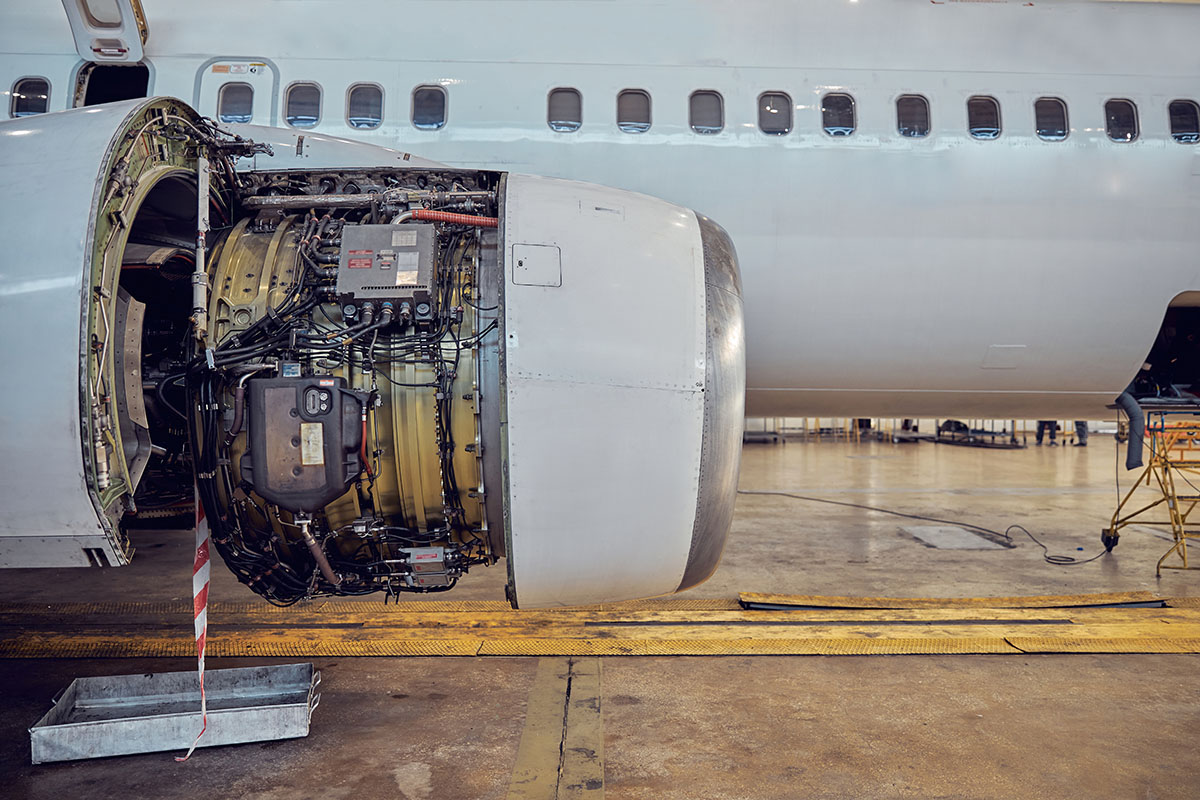 A jet engine with its cowling opened for maintenance, exposing wiring, pipework and accessory units, beneath the wing of an aircraft in a hangar