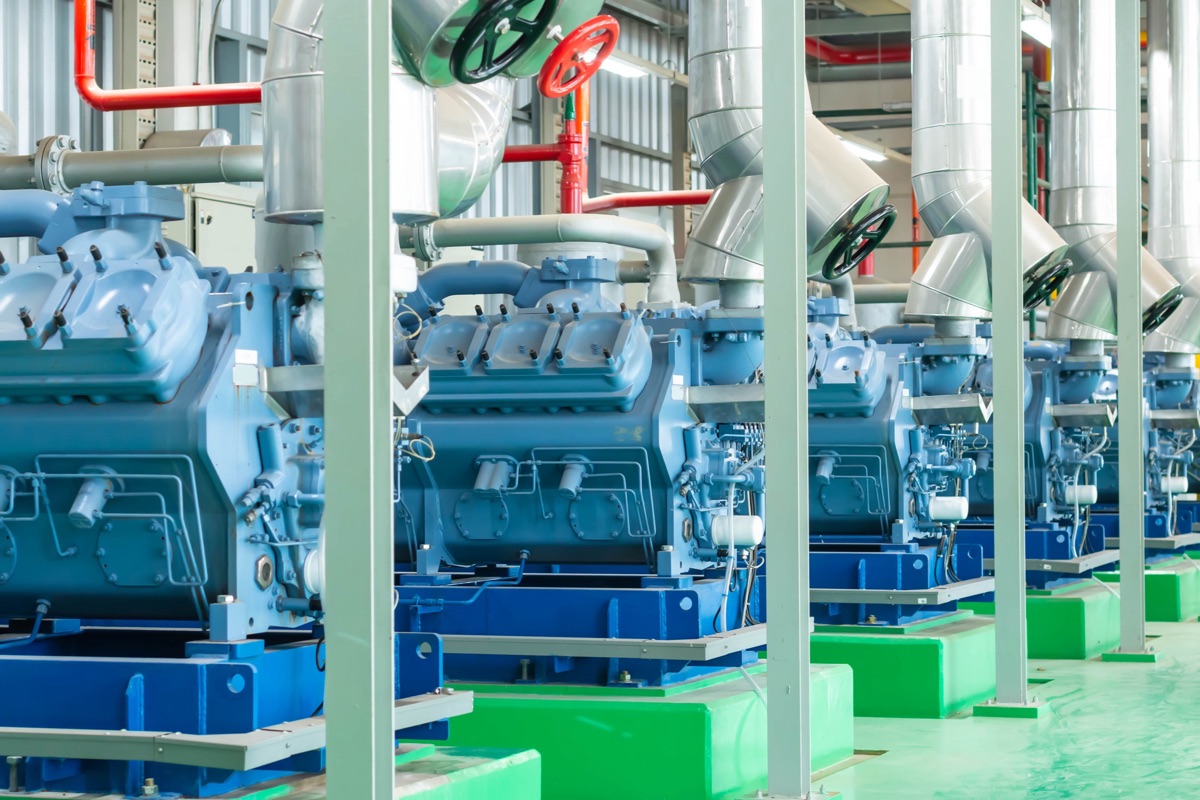 A row of blue industrial compressors on green painted plinths inside a plant machinery hall with insulated pipework overhead