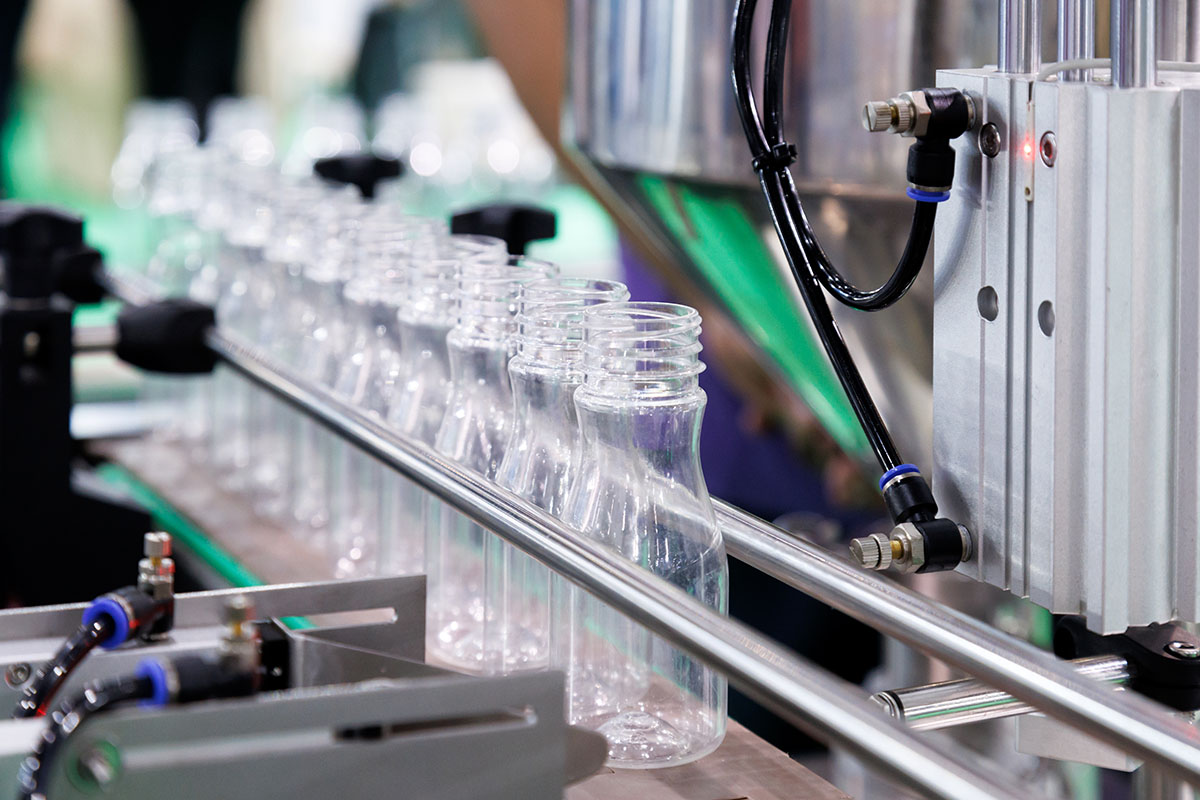 Rows of empty clear plastic bottles moving along a stainless steel conveyor rail into the pneumatic heads of an automatic filling machine