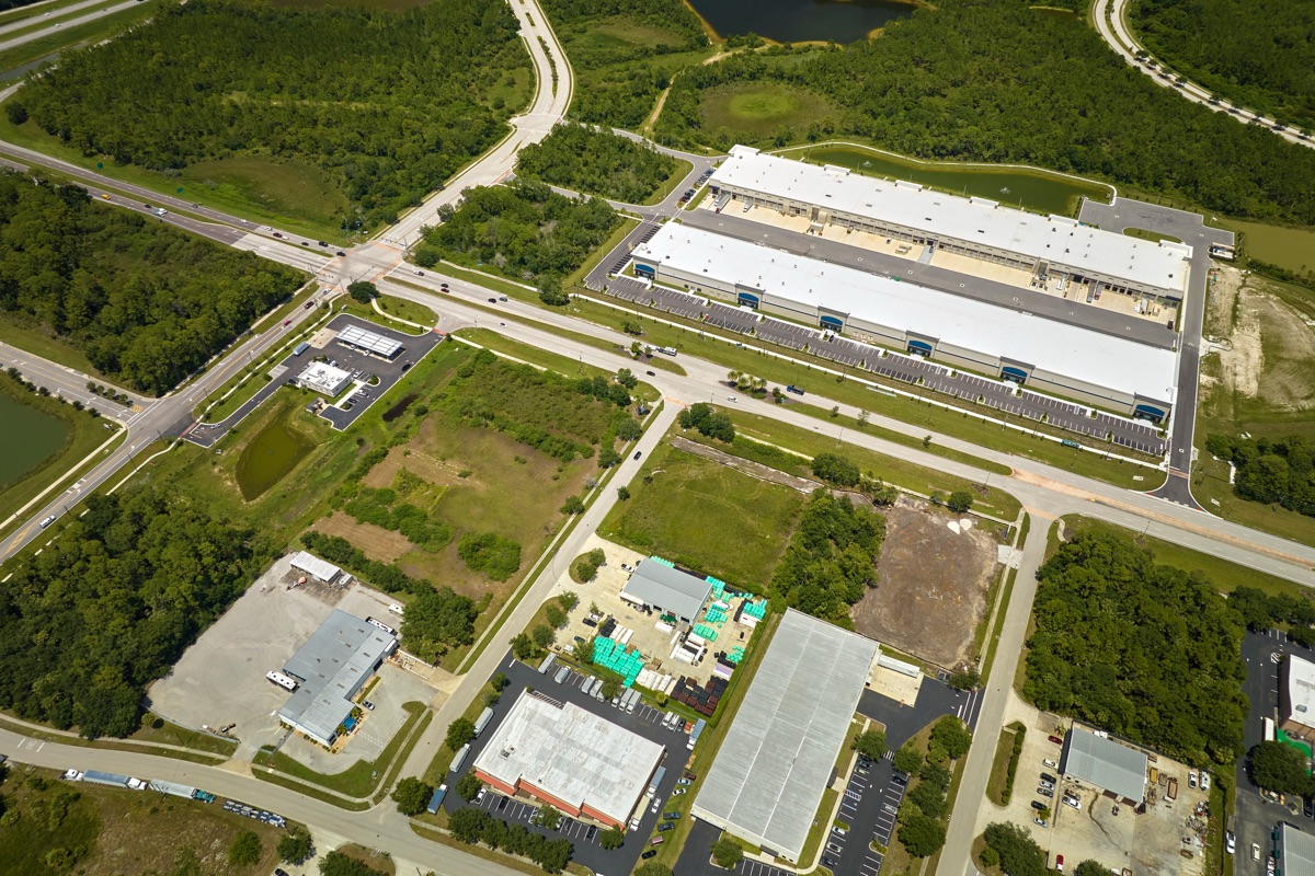 Aerial view of two large white-roofed distribution warehouses with loading docks and parking, set among roads and woodland in an industrial park