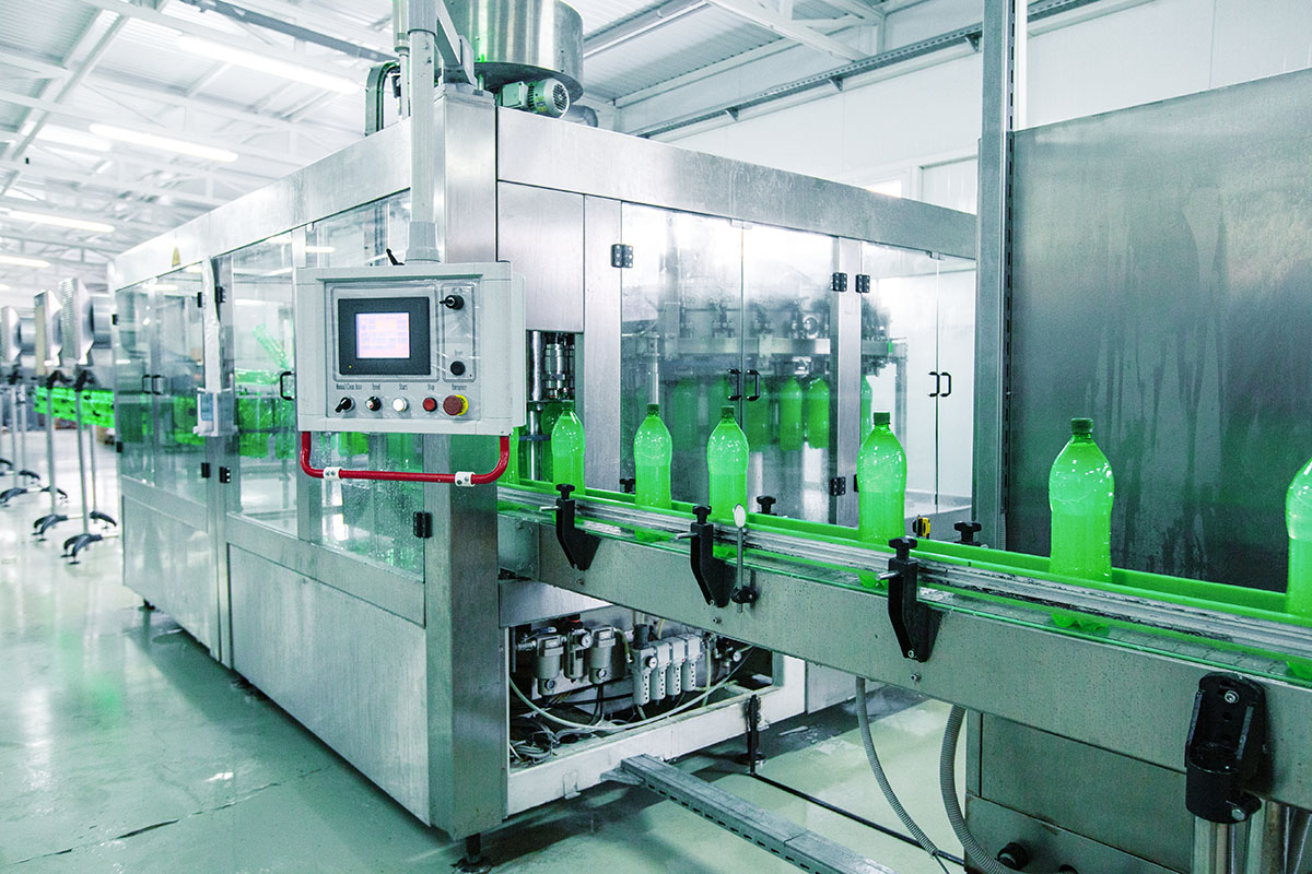 A stainless steel bottle filling machine with a touchscreen control panel feeds green plastic bottles along a conveyor in a bright beverage plant