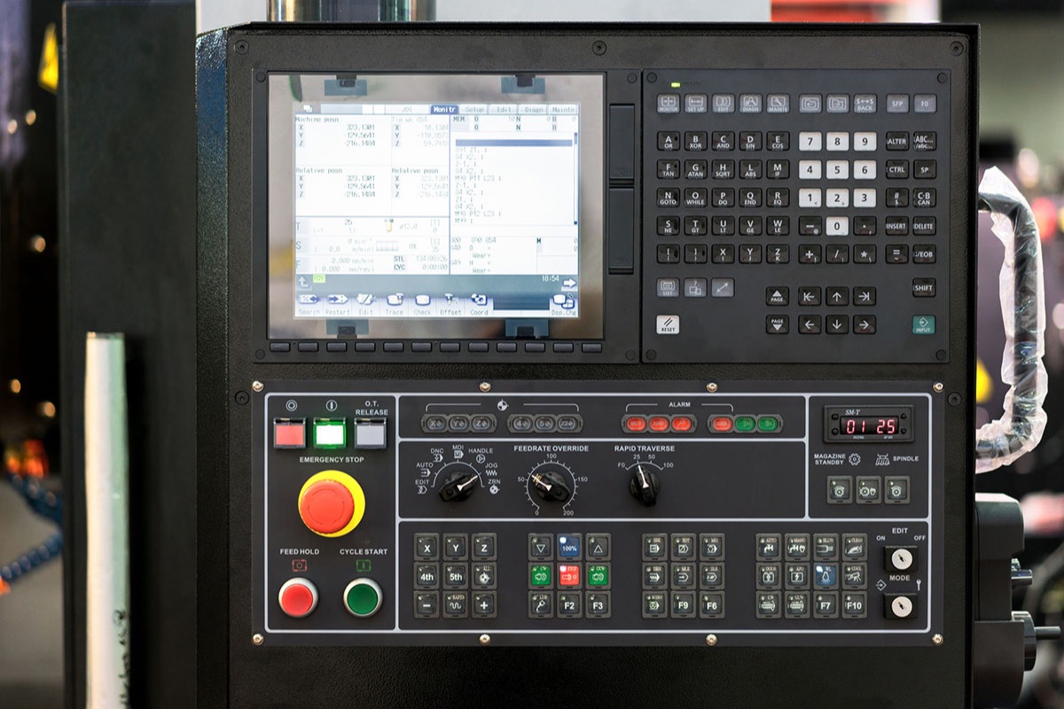 The control panel of a CNC machining centre, with a programming screen, keypad, emergency stop button and feed rate dials