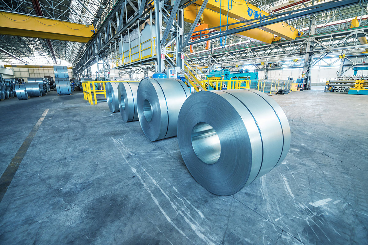 Coils of cold-rolled steel lined up on the concrete floor of a large metal processing plant beneath yellow overhead cranes