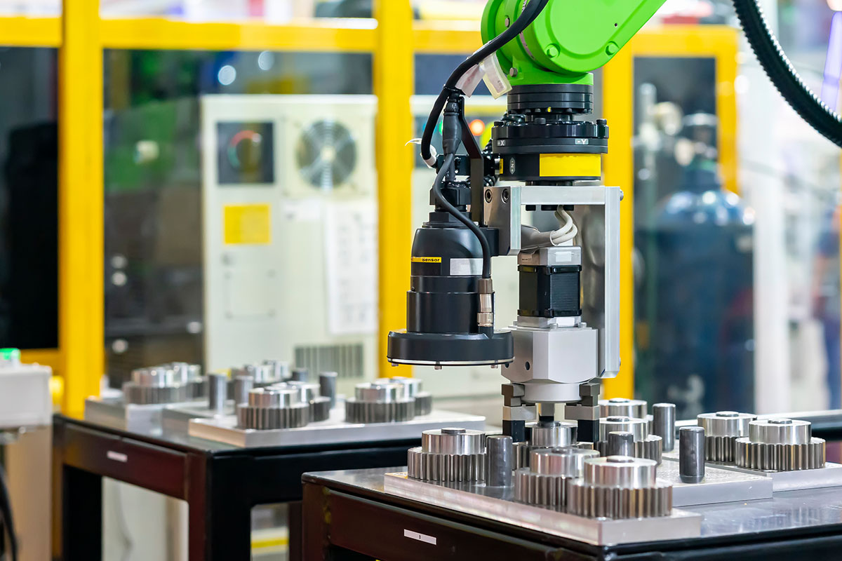 A green collaborative robot arm fitted with an inspection camera and gripper works over trays of machined steel gears on a production cell