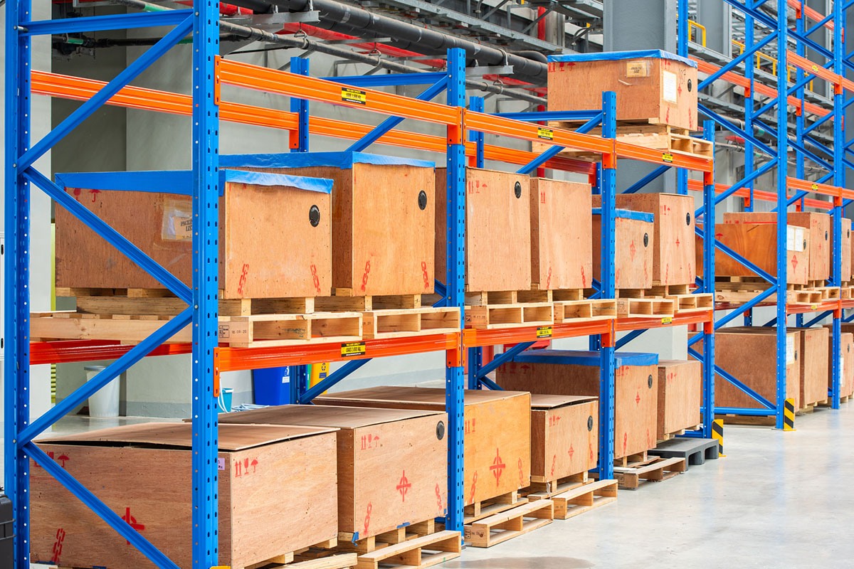 Wooden crates of boxed parts stacked on pallets across blue and orange steel racking in a clean industrial warehouse