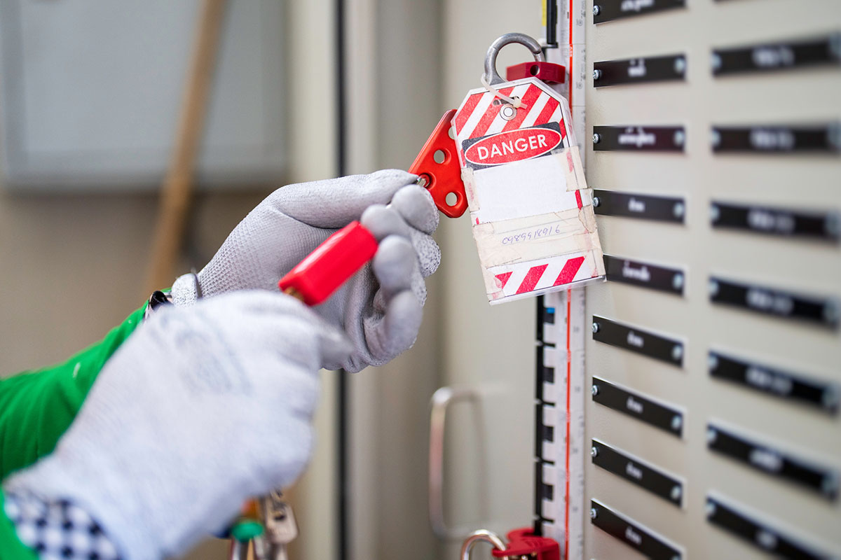 A technician in grey work gloves clips a red lockout hasp and a striped danger tag onto an electrical isolation panel