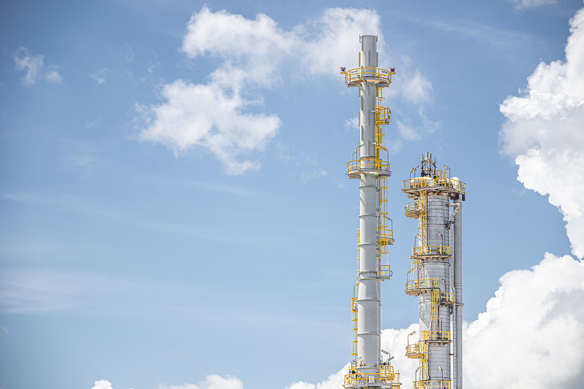 Two white refinery distillation columns with yellow ladders and platforms rising against a blue sky with white clouds