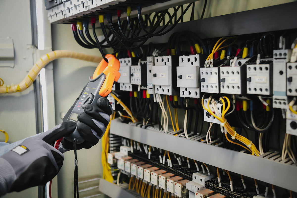 A gloved technician holds a clamp meter against the wiring of an industrial control cabinet lined with contactors and terminal blocks