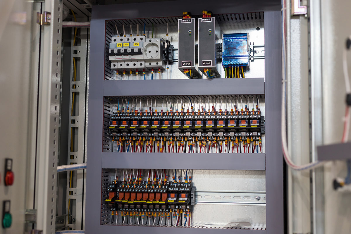 An open industrial control cabinet with power supplies, circuit breakers and rows of relays wired on DIN rails