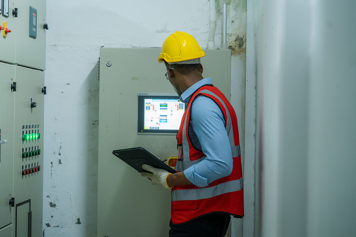 A technician in a hard hat and safety vest inspecting an HMI touchscreen on an industrial electrical control panel